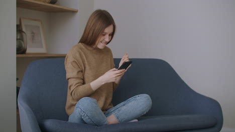 A-woman-in-writing-a-message-on-her-brand-new-smartphone-while-sitting-on-the-couch-with-a-white-pillow-at-home.